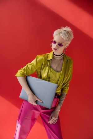 Blonde Albino-Frau mit Tätowierung posiert in trendiger Sonnenbrille und hält Laptop auf karminrosa 