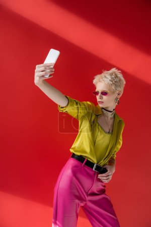 Foto de Mujer albina tatuada en gafas de sol elegantes tomando selfie en el teléfono inteligente sobre fondo rosa carmín - Imagen libre de derechos