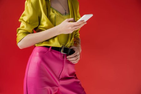 cropped view of tattooed woman in trendy outfit using smartphone on carmine pink background 