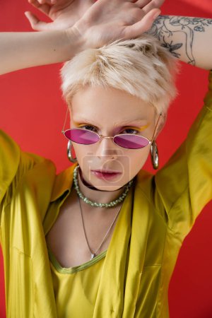 Porträt einer Albino-Frau mit Tätowierung posiert in stylischer Bluse und trendiger Sonnenbrille auf karminrosa 