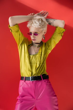 Blondine mit tätowierter Hand posiert in stylischer Bluse und trendiger Sonnenbrille auf karminrosa 