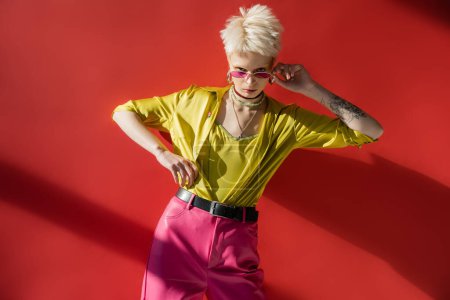 blonde albino woman with tattoo adjusting sunglasses on carmine pink background 