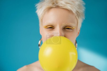blonde albino woman with bright eye liner blowing bubble gum on blue background 