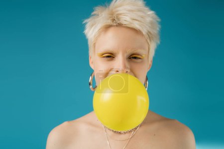 blonde albino woman with bare shoulders blowing bubble gum on blue background 