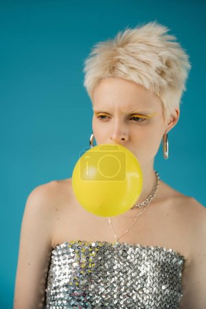 Téléchargez les photos : Élégant albinos femme avec les épaules nues soufflant gomme à bulles sur fond bleu - en image libre de droit