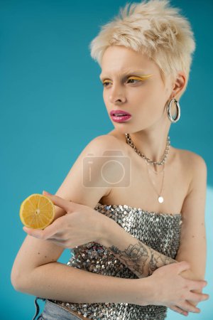 tattooed blonde albino woman with bare shoulders holding lemon half on blue 