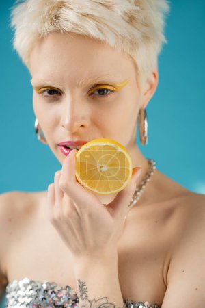 Blonde Albino-Frau mit nackten Schultern hält Zitronenhälfte auf Blau 