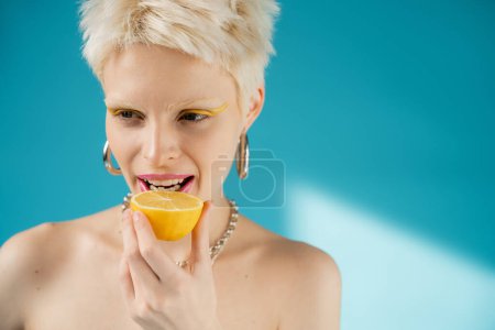 blonde albino woman with bare shoulders biting sour lemon half on blue background 