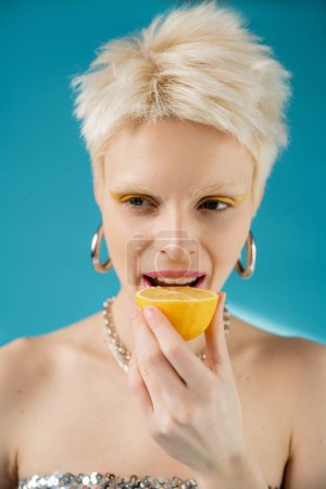 modelo albino rubia con hombros desnudos mordiendo medio limón agrio sobre fondo azul 