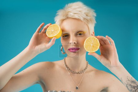 modelo albino rubia con tatuaje en la mano sosteniendo mitades de limón agrio sobre fondo azul 