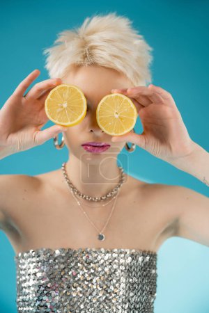 blonde albino model with bare shoulders covering face with lemon halves on blue 