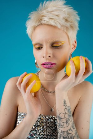 modèle albinos tatoué avec maquillage tendance et épaules nues posant avec des citrons sur bleu 