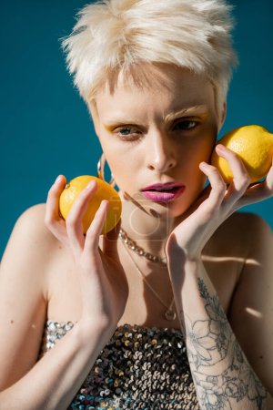 modèle albinos tatoué avec maquillage à la mode et cheveux blonds posant avec des citrons sur bleu 