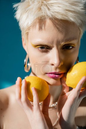 Porträt einer Albino-Frau mit trendigem Make-up und blonden Haaren, die mit reifen Zitronen auf Blau posiert 