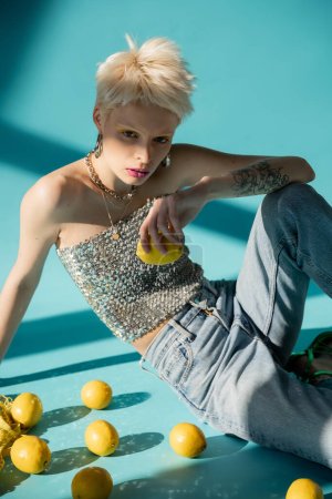 tattooed albino woman in shiny top with sequins sitting near ripe lemons on blue