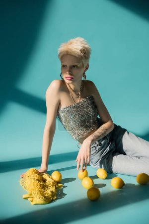 tattooed albino model in shiny top with sequins and jeans sitting near ripe lemons on blue 