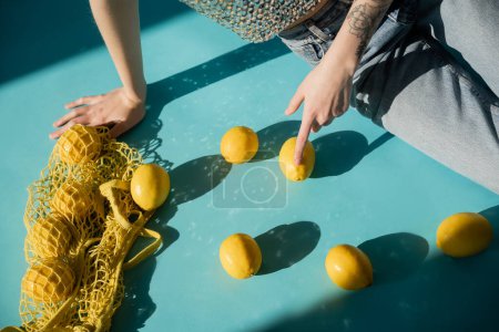 cropped view of tattooed woman in shiny top with sequins and jeans sitting near string bag with ripe lemons on blue 