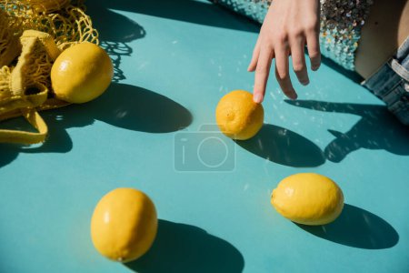 cropped view of woman in shiny top with sequins posing near string bag and ripe lemons on blue 