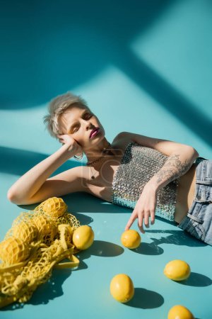 Photo for Tattooed albino model in shiny top with sequins and jeans posing near ripe lemons on blue - Royalty Free Image