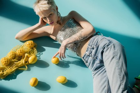 overhead view of tattooed albino woman in top with sequins and jeans lying near ripe lemons on blue 