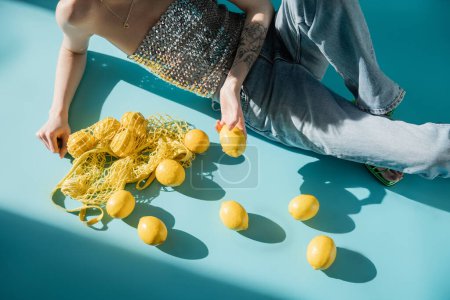 high angle view of tattooed woman in shiny top with sequins and jeans sitting near string bag and ripe lemons on blue 