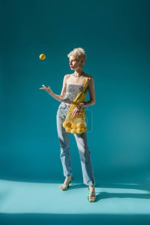 full length view of tattooed woman in shiny top with sequins holding net bag and throwing in air ripe lemon on blue 