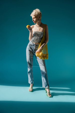 full length view of tattooed woman in shiny top with sequins holding string bag with ripe lemons on blue 