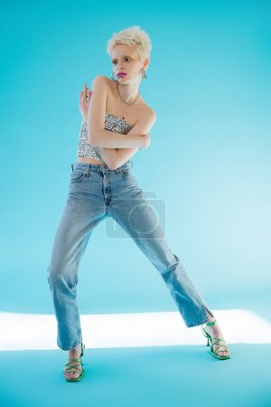 full length view of tattooed albino model in shiny top with sequins and denim jeans posing on blue 