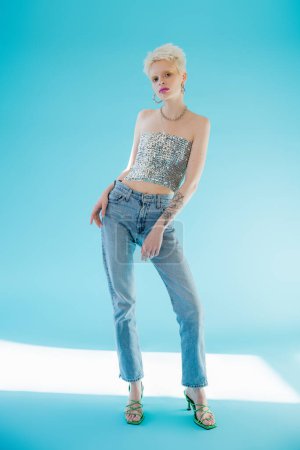 full length view of pretty albino model in shiny top with sequins and denim jeans posing on blue 
