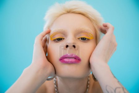 Photo for Blonde albino model with bright makeup looking at camera on blue background - Royalty Free Image