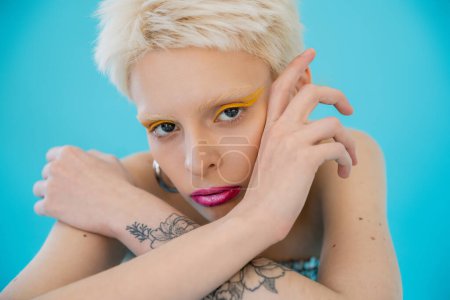 tattooed and blonde albino model with bright makeup looking at camera on blue background 