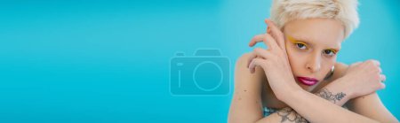 Photo for Tattooed and blonde albino model with bright makeup looking at camera on blue background, banner - Royalty Free Image