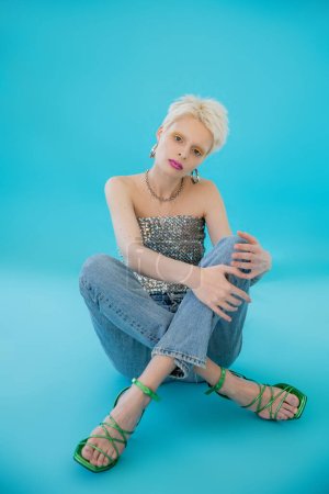 full length of blonde albino woman with heeled sandals and jeans sitting on blue 