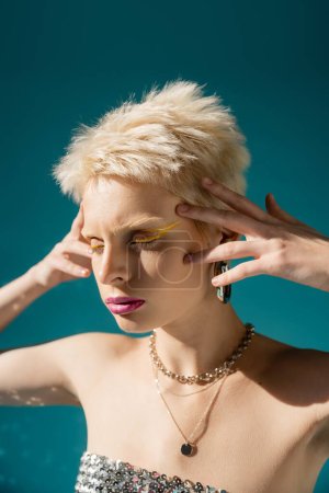 retrato de mujer albina rubia con maquillaje de moda posando con las manos cerca de la cabeza en azul 