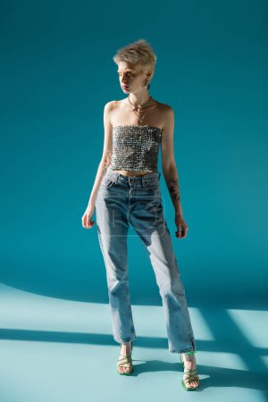 full length view of tattooed albino model in top with sequins and denim jeans standing on blue 