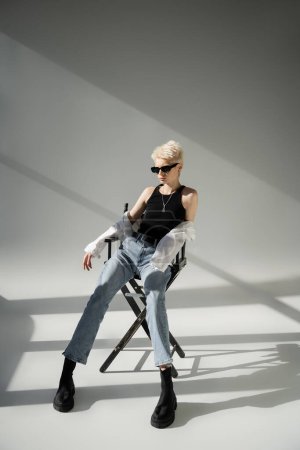 volle Länge der stilvollen blonden Frau in Sonnenbrille und trendigem Outfit sitzt auf Klappstuhl auf grau 