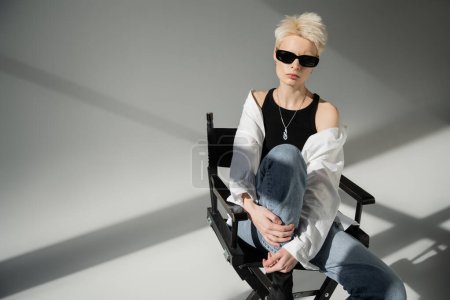 stylische blonde Frau mit schwarzer Sonnenbrille und trendigem Outfit sitzt auf Klappstuhl auf grauem Hintergrund 