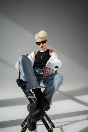 volle Länge der stilvollen blonden Frau in trendiger Sonnenbrille und schwarzen Stiefeln sitzt auf Klappstuhl auf grau 