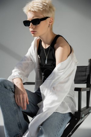 blonde woman in trendy sunglasses and stylish clothes sitting on folding chair on grey 