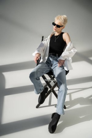 pleine longueur de femme blonde élégante en lunettes de soleil noires assis sur chaise pliante sur gris 