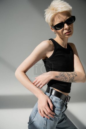 stylish blonde woman with tattoo posing in black sunglasses on grey 