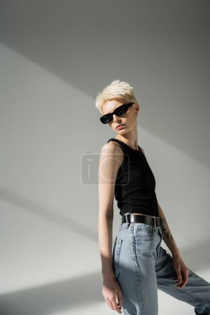 stylish blonde woman in tank top and jeans posing in black sunglasses on grey background 