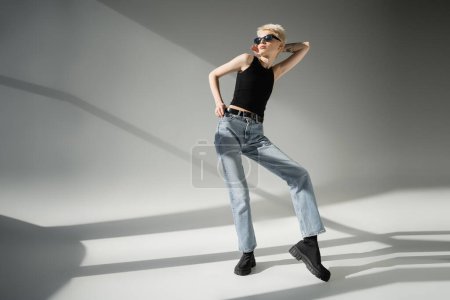full length of tattooed model in black sunglasses and tank top posing on grey background 