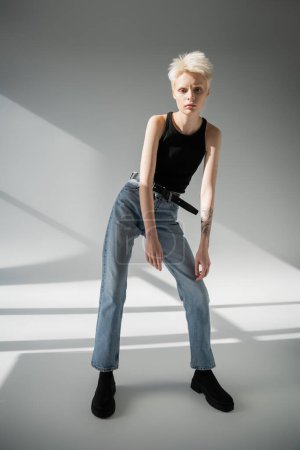 full length of young albino woman with tattoo on hand posing in jeans and tank top on grey 