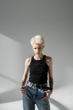 ernsthafte Albino-Frau mit Tätowierung auf der Hand posiert in Jeans und Tank-Top auf grau 