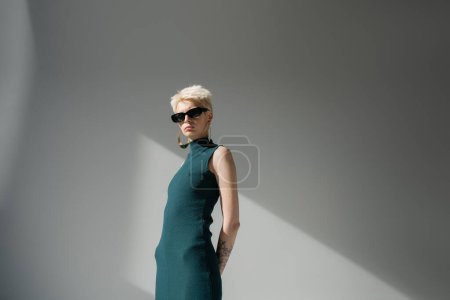 tätowiertes Model mit heller Haut posiert in stylischer Sonnenbrille und Kleid auf grauem Hintergrund 