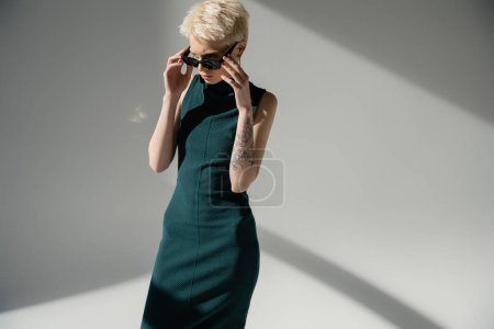 blonde woman with fair-skin and tattoo adjusting stylish sunglasses on grey background 