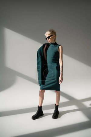 Photo for Full length of tattooed and blonde woman in sunglasses posing in green dress and coat on grey background - Royalty Free Image