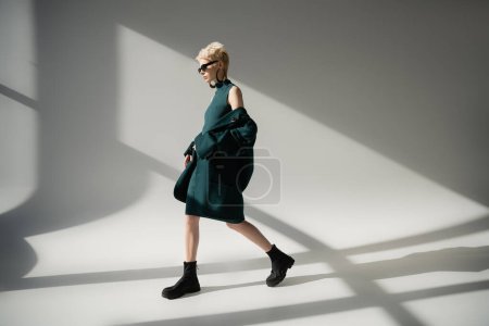 full length of blonde woman with fair-skin walking in dress and coat on grey background 