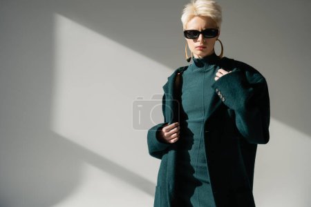 young woman in trendy sunglasses standing in green dress and coat on grey background 